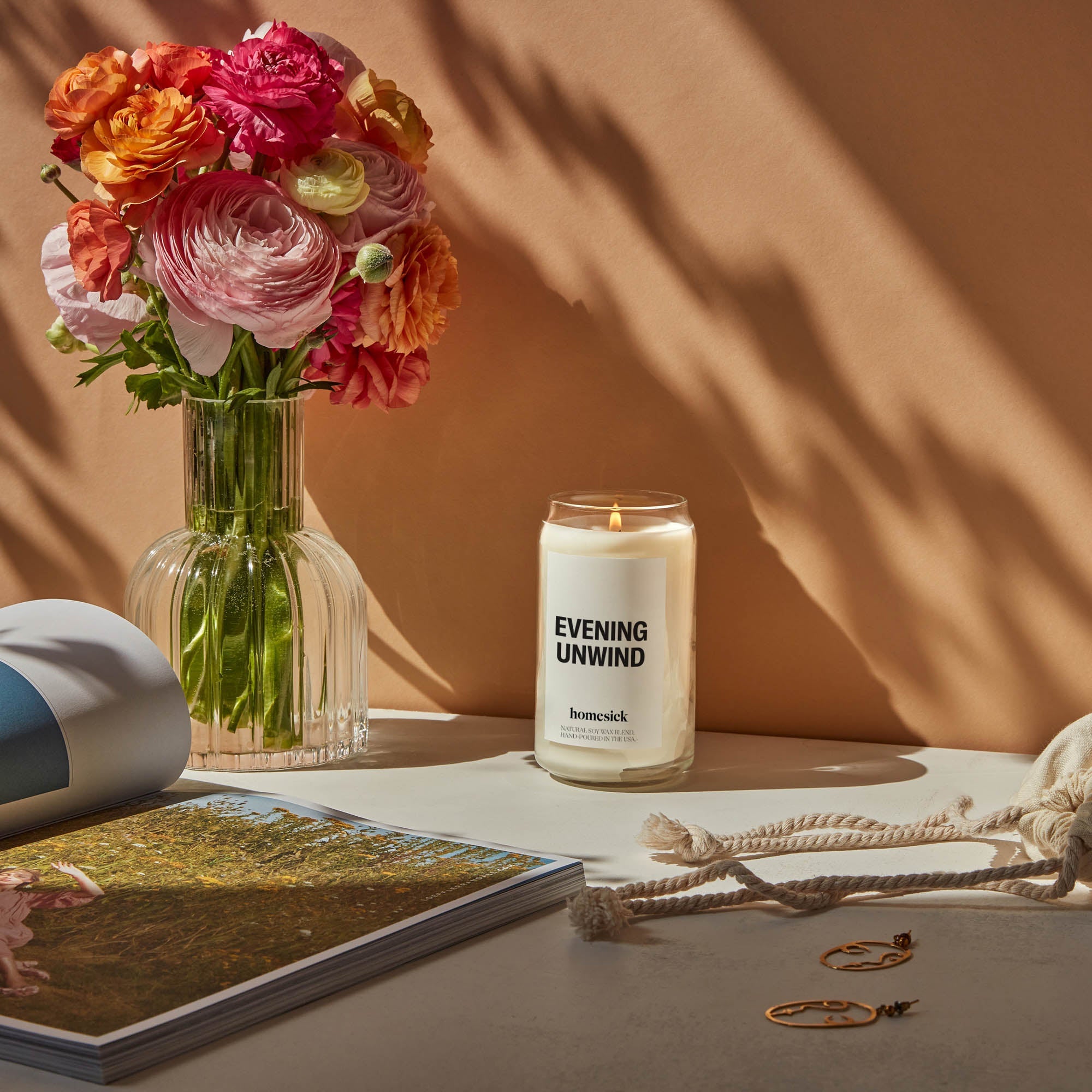 Lit evening unwind candle with a vase of flowers on a dresser with light coming through a window.