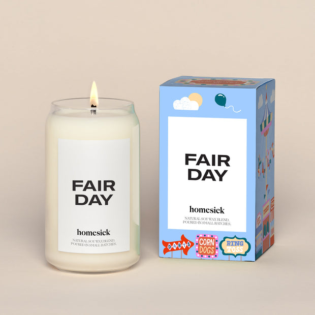 A lit Fair Day Homesick candle displayed next to its boxed packaging on a dark cream background.