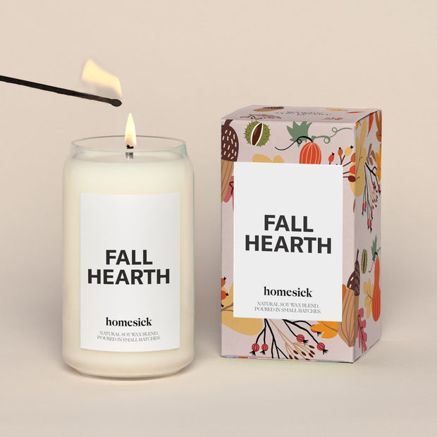 A lit Fall Hearth Homesick candle displayed next to its boxed packaging on a dark cream background.