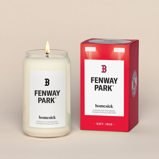 A lit Fenway Park Homesick candle displayed next to its boxed packaging on a dark cream background.