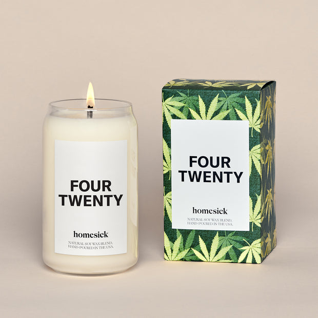 A lit Four Twenty Homesick candle displayed next to its boxed packaging on a dark cream background.