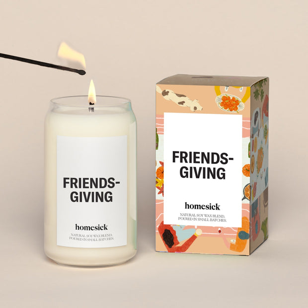 A lit Friendsgiving Homesick candle displayed next to its boxed packaging on a dark cream background.
