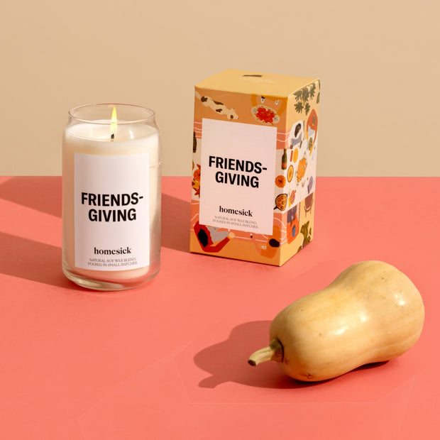 The Friendsgiving Candle and its packaging shot on a salmon surface with a dark cream background. There is a gourd in front of the candle.