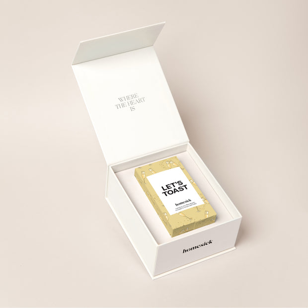 A white candle box that displays how a candle fits perfectly inside. The top lid packaging reads, "where the heart is" and inside is the Let's Toast candle. 