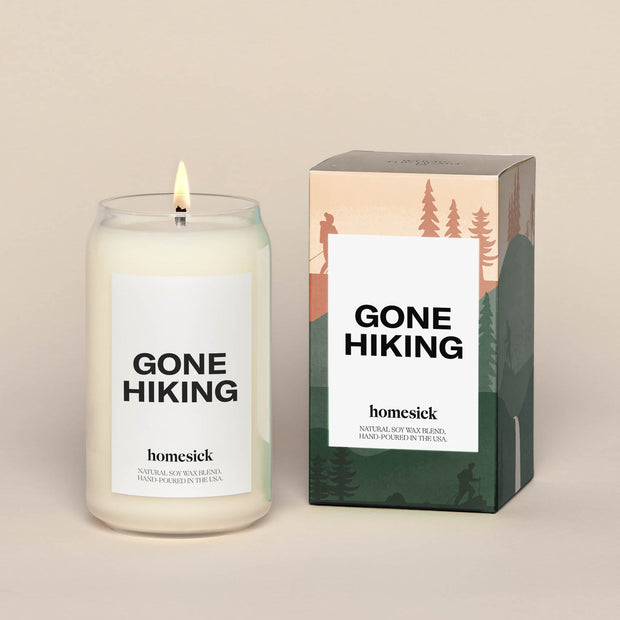 A lit Gone Hiking Homesick candle displayed next to its boxed packaging on a dark cream background.