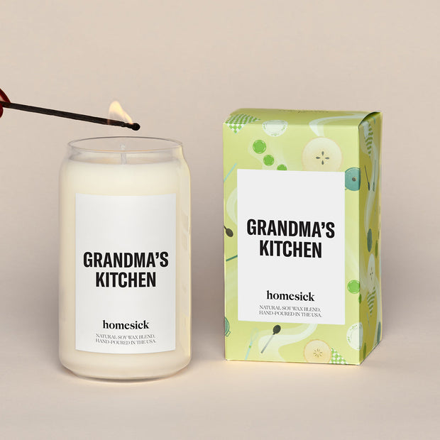 A lit Grandma's Kitchen Homesick candle displayed next to its boxed packaging on a dark cream background.