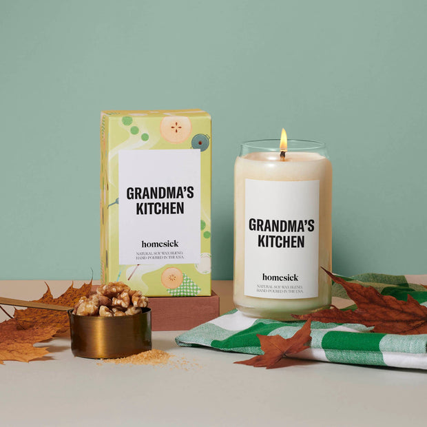 A stylized shot of the lit Grandma's Kitchen candle next to its packaging. The gray surface has a cup of walnuts and leaves with a mint green background.