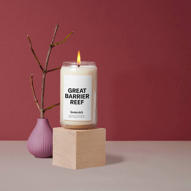 A Great Barrier Reef Candle displayed on top of a wooden cube pedestal. Next to it, is a deep lilac container with a branch. The surface is a medium gray color and wine background.