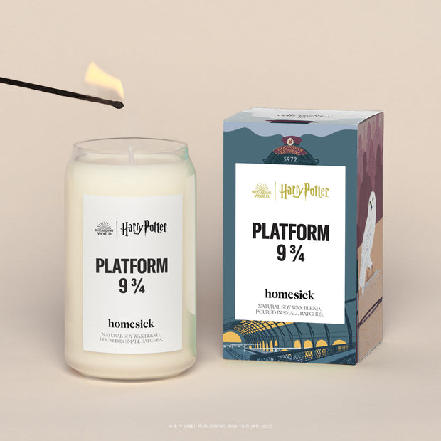 A lit Platform 3/4 Homesick candle displayed next to its boxed packaging on a dark cream background.