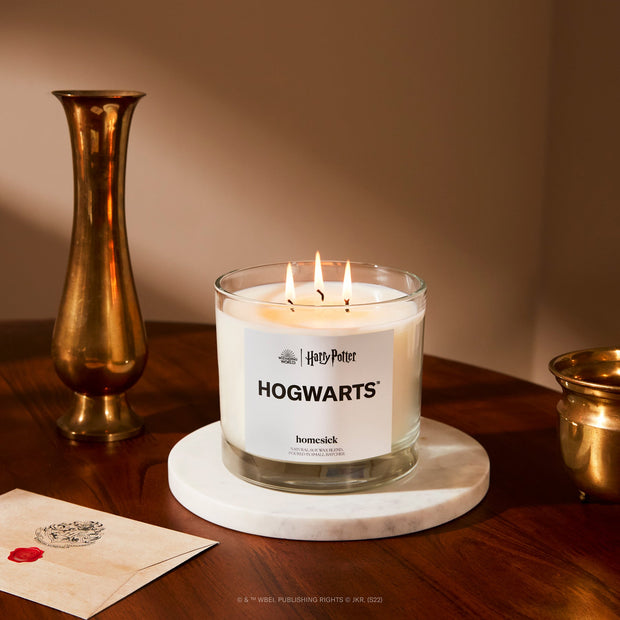 A lifestyle image of the 3-Wick Hogwarts candle displayed on a round white marble coaster. The coaster sits on a dark wood table with gold props also on it. 