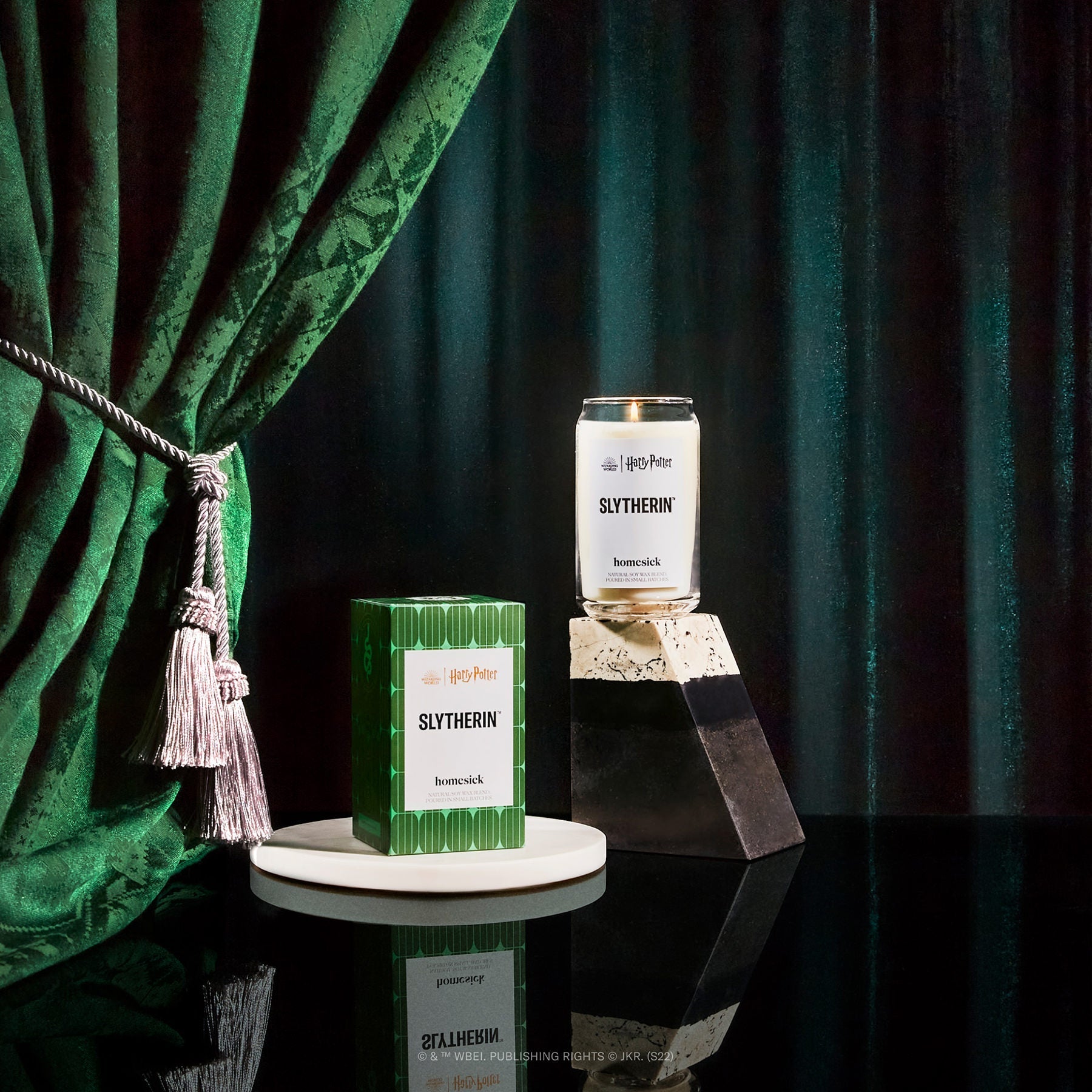A product shot of the Slytherin candle on a dark glossy surface. There is a green curtain and fabric for the background.