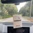 The Hawaii Car Freshener hanging on a rear view mirror. One can see out the road ahead with various greenery on the sides.