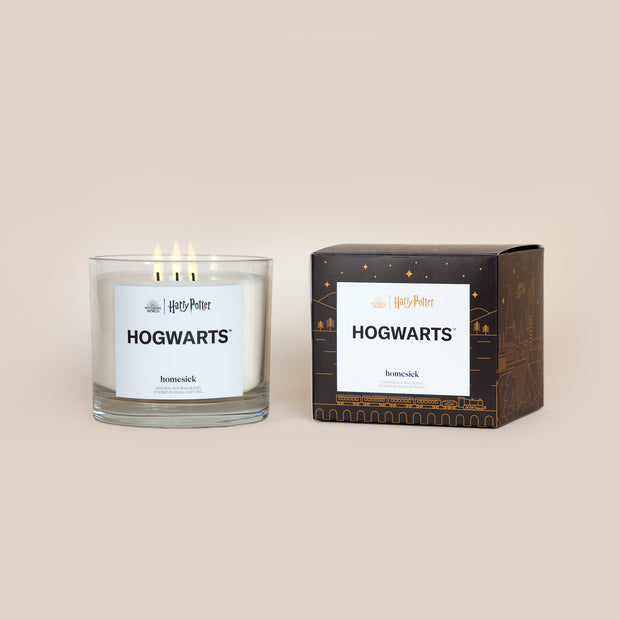 A lit 3-Wick Hogwarts Homesick candle displayed next to its boxed packaging on a dark cream background.