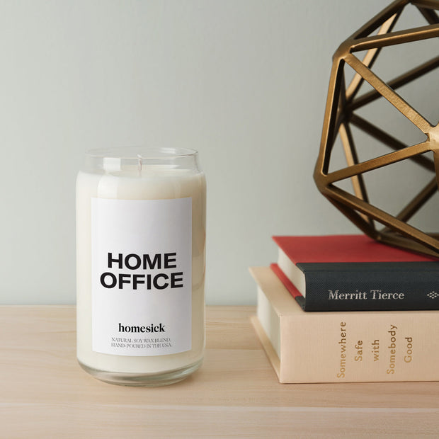 A close up of the Home Office Candle that is on display on a light wood surface. Next to the candle is a stack of books.