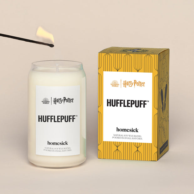 A lit Hufflepuff Homesick candle displayed next to its boxed packaging on a dark cream background.