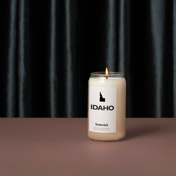 An elevated product shot of the Idaho Homesick candle. It is display on a luxurious gray surface with a silky black curtain as the background.