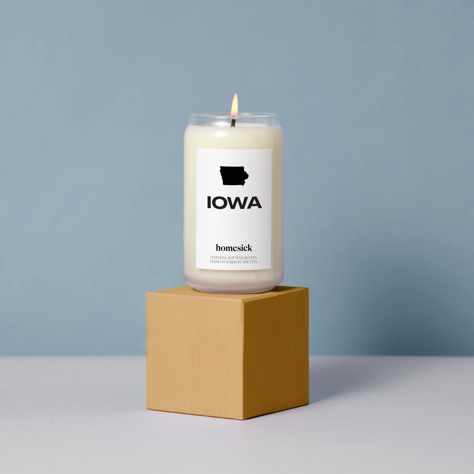 A Iowa Homesick candle placed on top of a dark yellow cube pedestal. The pedestal is in the middle of a gray surface and light blue background.