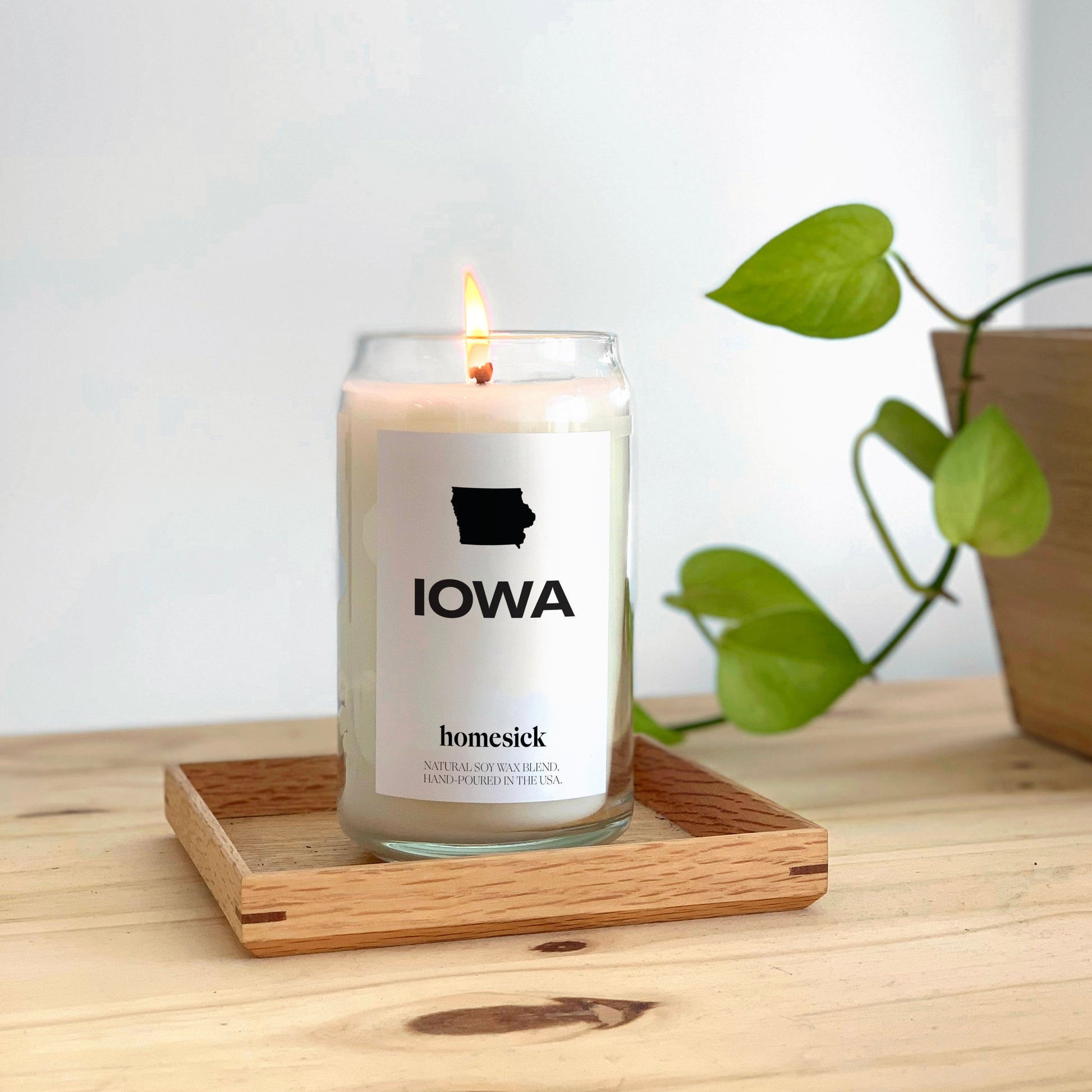 A closeup of the Iowa homesick candle that is placed inside a square tray. The tray is on top of a wooden surface and white background.
