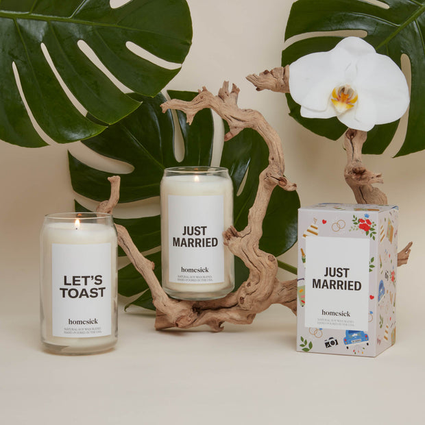 A group shot of the celebratory candles available at Homesick. Left to right there is the Let's Toast Candle, Just Married candle and the packaging for Just Married. Behind the products are palm leaves and a wooden branch.