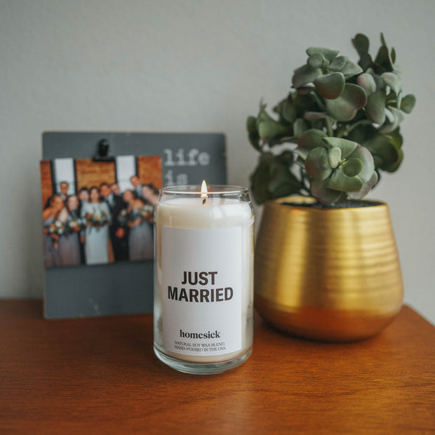A close up of the Just Married Candle on a wooden table. The background is blurred but there is a photo of a bridal party on the left and a plant in a gold pot to the right.