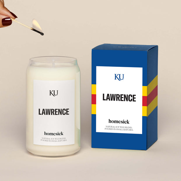 A lit Lawrence Homesick candle displayed next to its boxed packaging on a dark cream background.