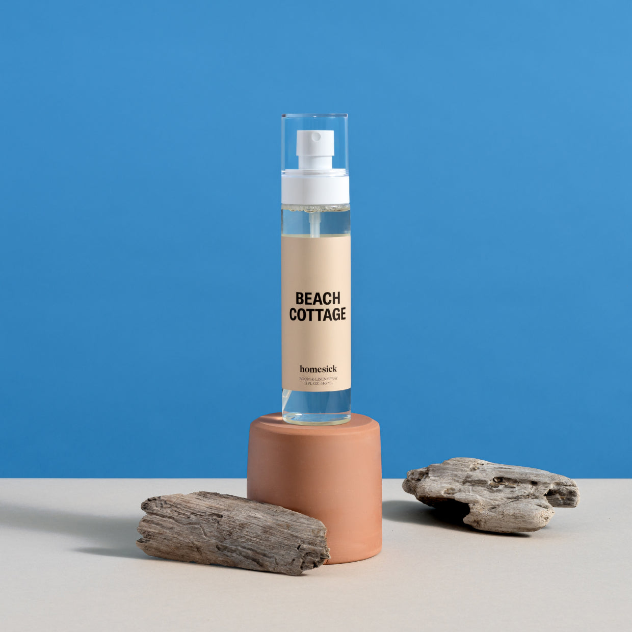 A bottle of Beach Cottage Room Spray displayed on top of a cup with coastal props on the table surface with a blue background.