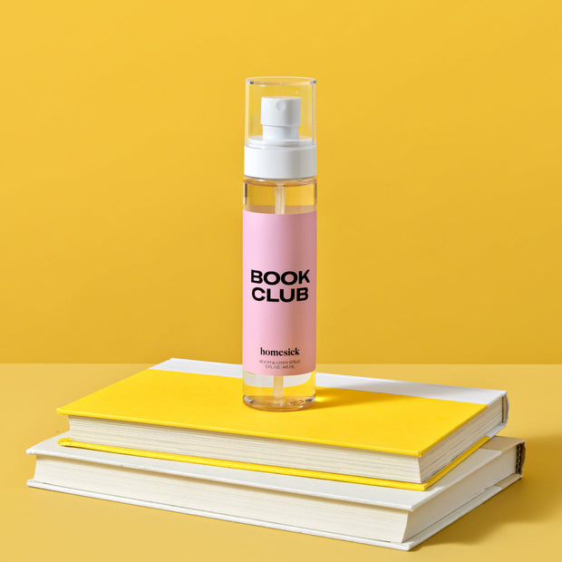 A bottle of Book Club Room Spray on a top of yellow books and there is a bright yellow background.