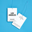 The front and the back of two Los Angeles Car Freshener tags. Both are shot overhead on a bright blue background.