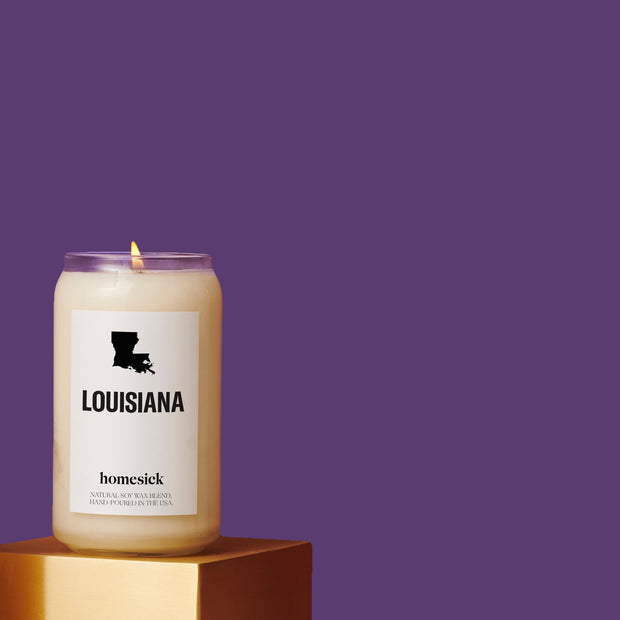 A Louisiana Homesick candle on a golden pedestal in the lower left of the image. The rest is negative space of a dark purple background.
