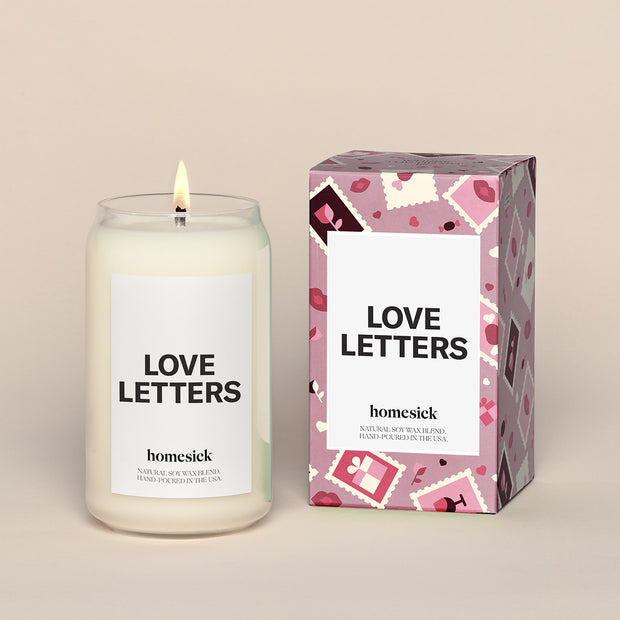 A lit Love Letters Homesick candle displayed next to its boxed packaging on a dark cream background.