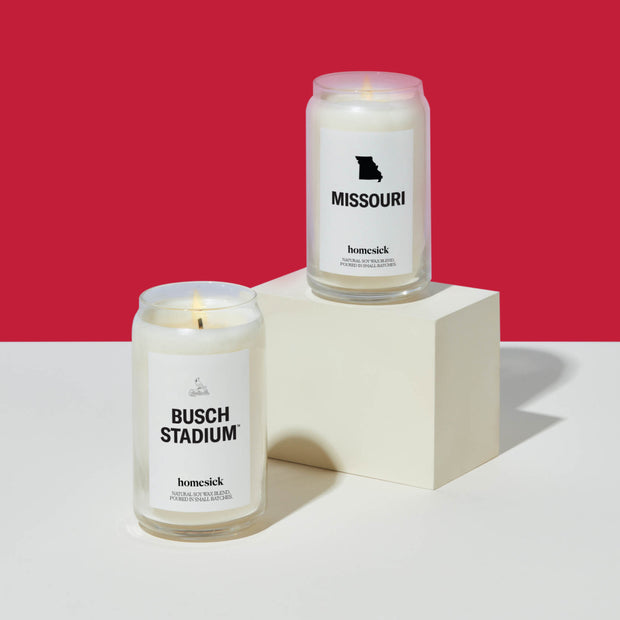 Two lit Homesick candle displayed next to its boxed packaging on a dark cream background.