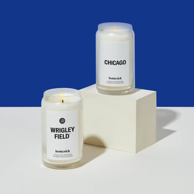 A Wrigley Field Homesick candle on top of a white surface. To the right of this candle is the Chicago Homesick candle that sits upon a square pedestal. Both are in front of a blue background.