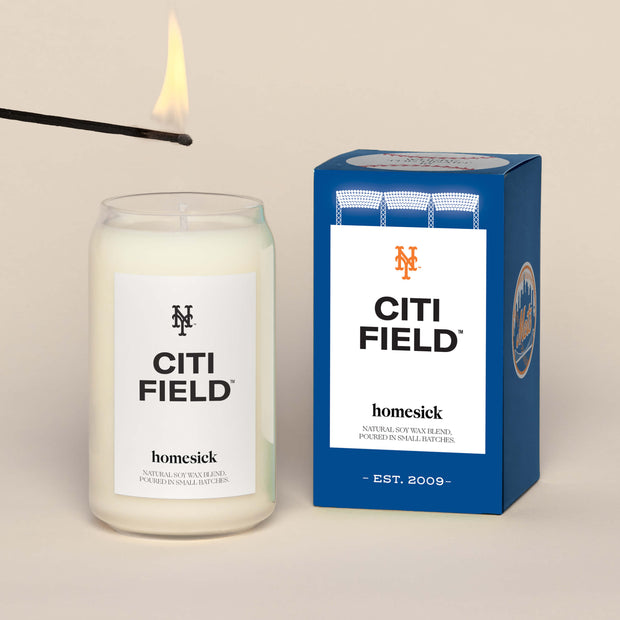 A lit Citi Field Homesick candle displayed next to its boxed packaging on a dark cream background.