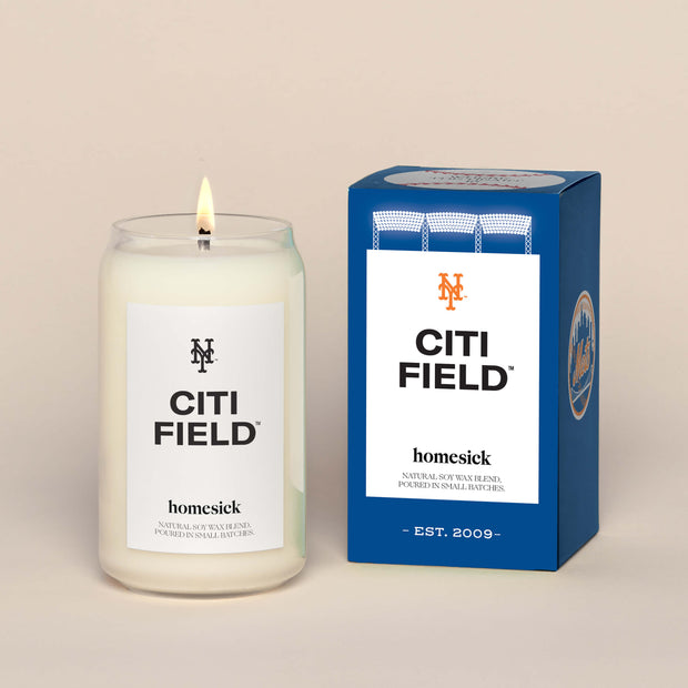 A lit Citi Field Homesick candle displayed next to its boxed packaging on a dark cream background.