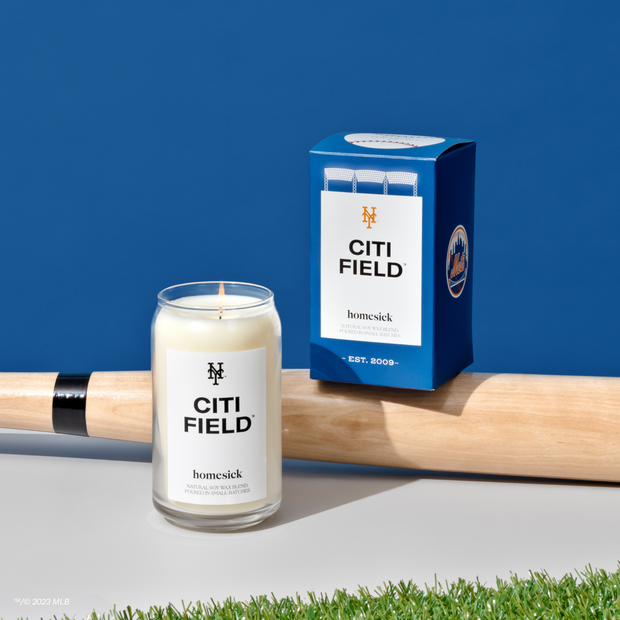 A Citi Field Candle displayed on a light gray surface. A baseball bat is behind that candle and the Citi Field box is perched on top of the baseball.