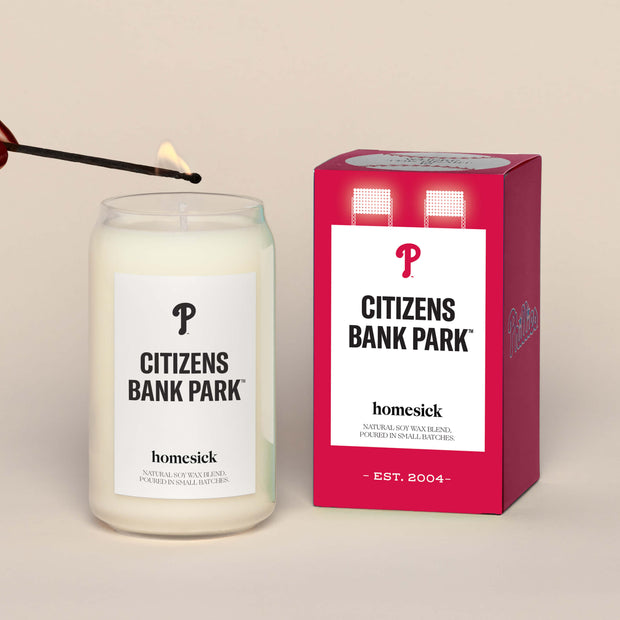 A lit Citizens Bank Park Homesick candle displayed next to its boxed packaging on a dark cream background.