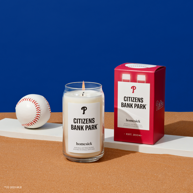 A Citizens Bank Park candle on a orangish surface with a blue background. Next to the candle itself is a baseball and the packaging for the candle.