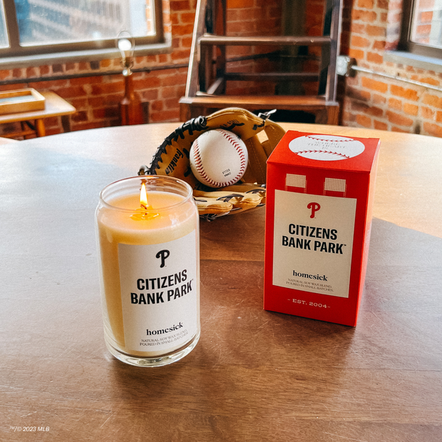 A lifestyle image of the Citizen Bank Park candle next to its packaging with a baseball and mit behind those.
