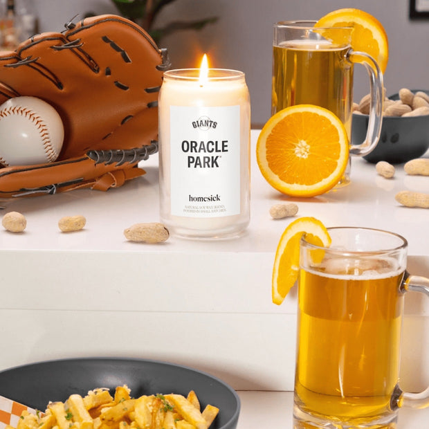 The Oracle Park Candle displayed on a glossy white surface with various baseball inspired props around it: baselball glove and ball, beer and orange slices.