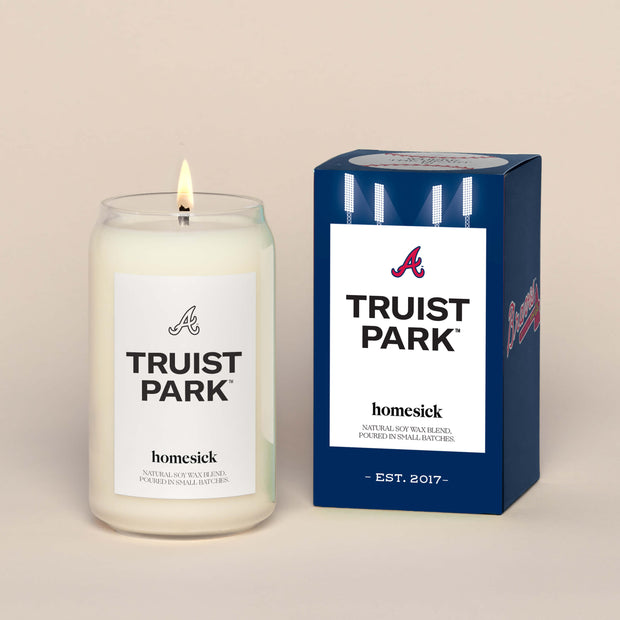 A lit Truist Park Homesick candle displayed next to its boxed packaging on a dark cream background.