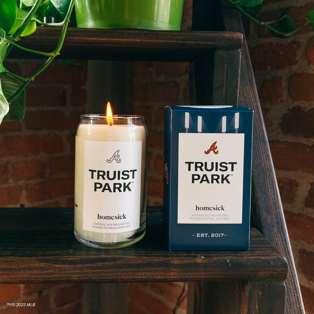 A lit Truist Park Candle next to its boxed packaging on display on a wooden shelf. One can see a green plant on the shelf above it and the brick wall around it.
