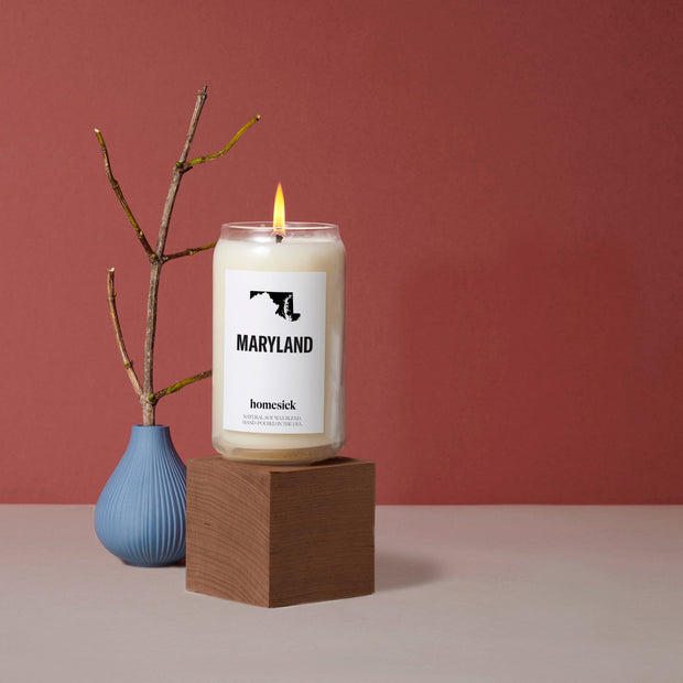 A Maryland Candle displayed on top of a wooden cube next to a small blue decor vase that has a dried branch in it. The surface is dark gray with a deep red background.