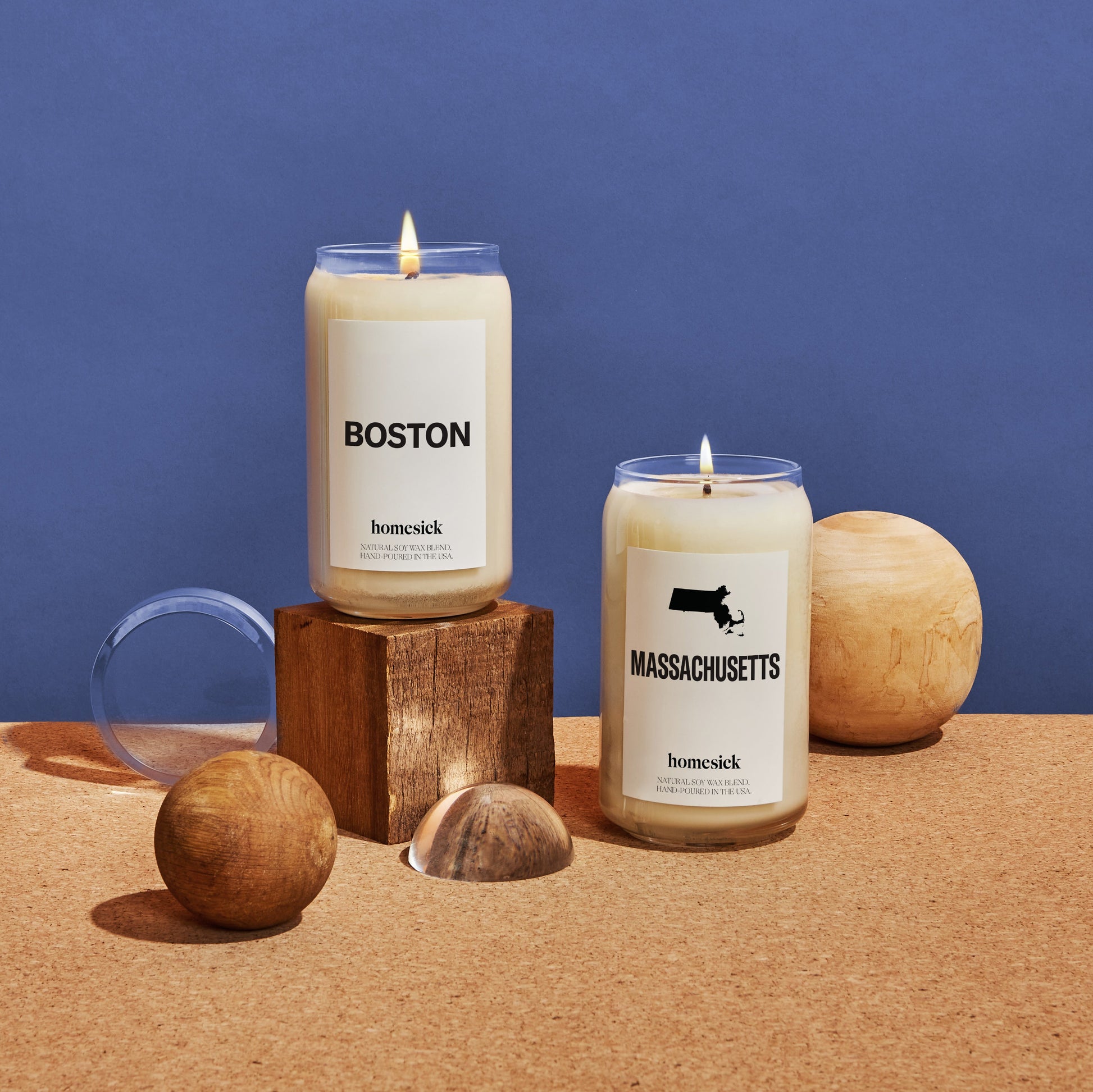 Two Homesick candles shot on a corkboard surface with various sphere and cube objects around them. The two candles shown are the Boston and Massachusetts candles in front of a blue background.