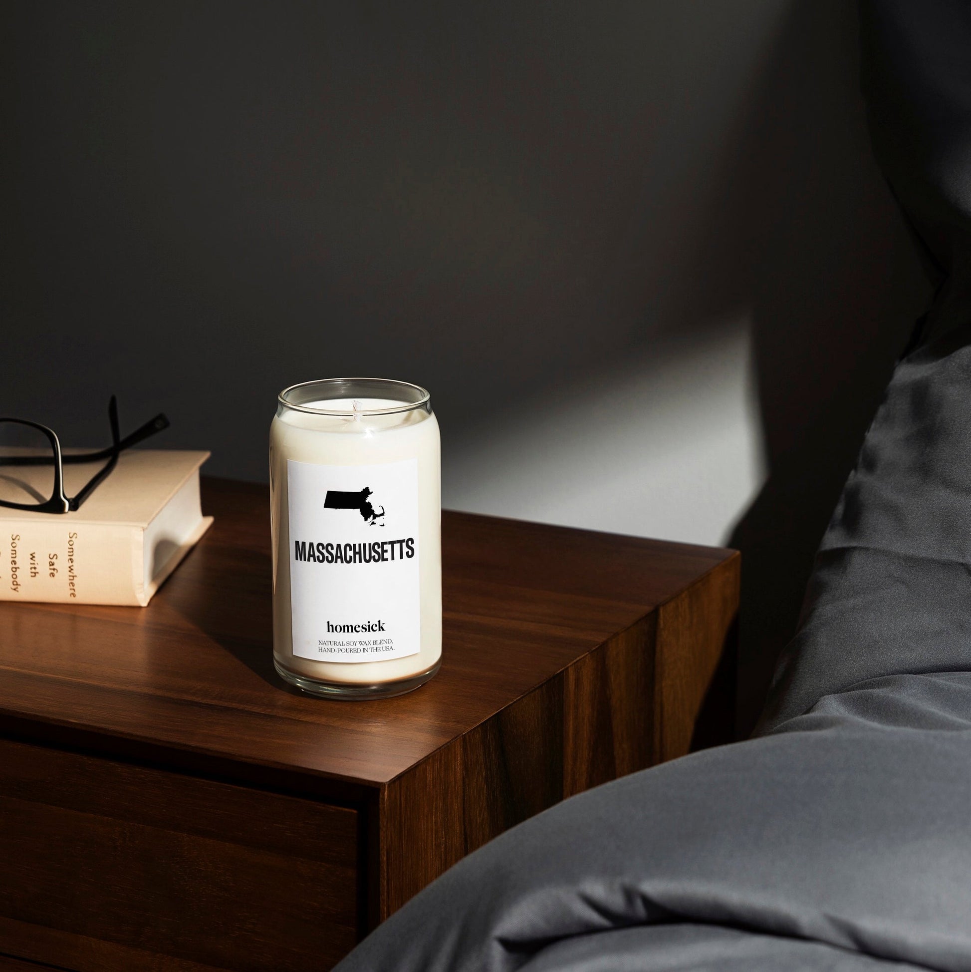 A bedside shot of the Massachusetts Candle on a deep glossy wood side table. One can see a cream book in the corner and the silky gray sheets of the bed.