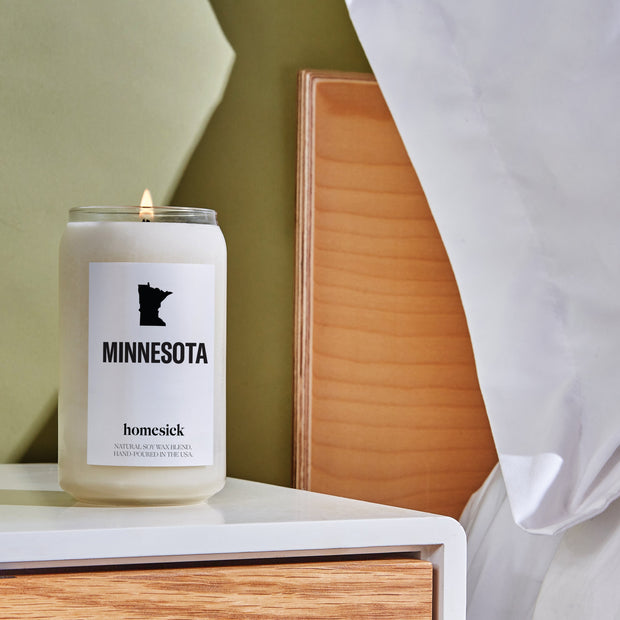 A bedside shot of the Minnesota candle on a white side table that has a wooden drawer.