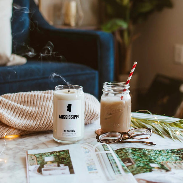 A lifestyle shot inside someone's living room where there is a recently blown out Mississippi Candle next to an iced coffee in a mason jar. Those items are on a white marble table with other personal items like a blanket and magazine.