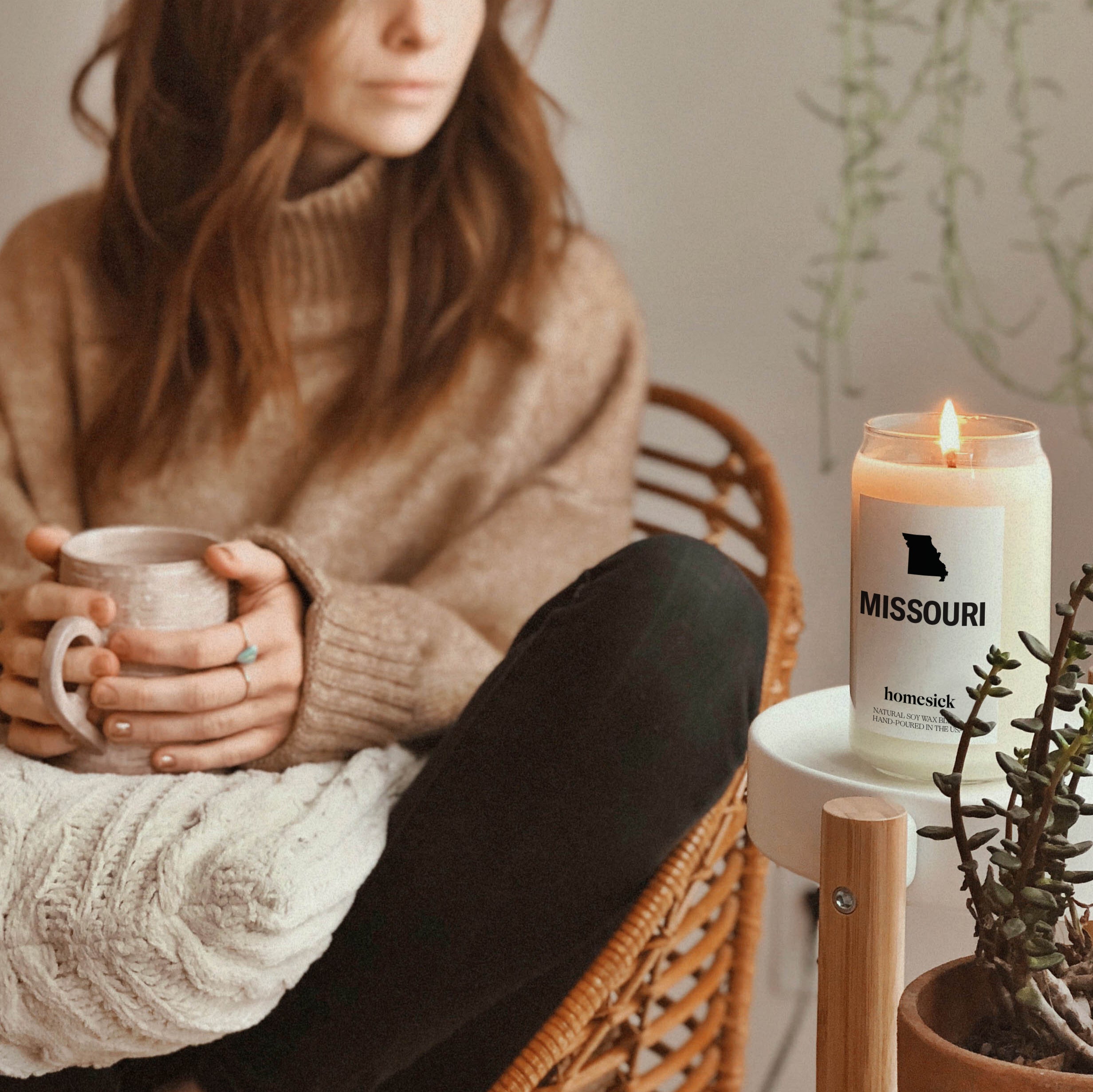 On the left is a woman who is holding a cup of tea sitting in a chair, her presence in the background is blurred. In the front corner is a Missouri candle lit on a white round sitde table.