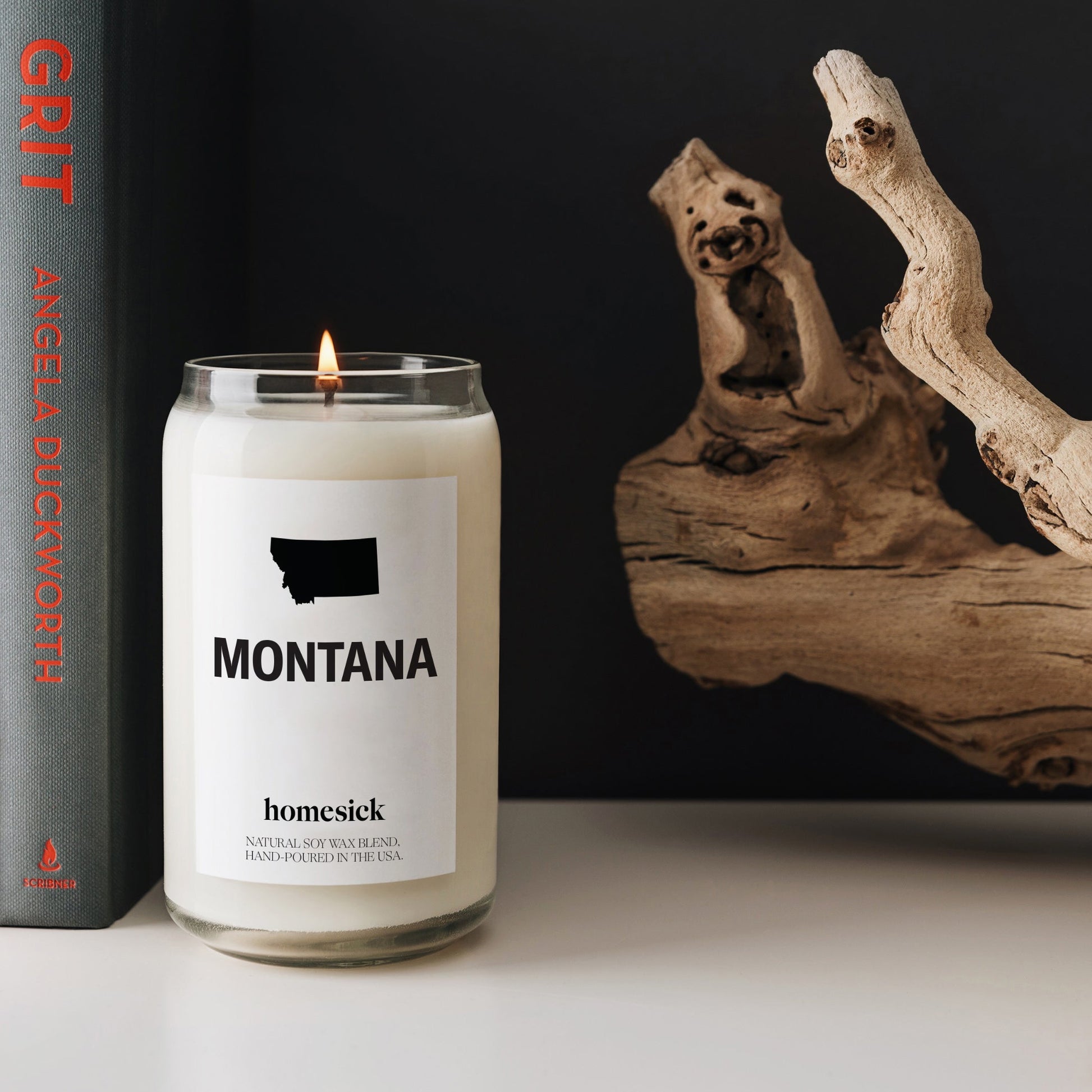 A close up of the Montana candle displayed on a white surface. Next to it is Angela Duckworth's book and to the right is a coastal log.