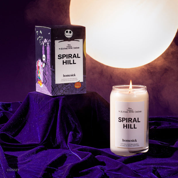 The Spiral Hill Candle on a purple velvet fabric surface. The boxed packaging is displayed on covered pedestal with a bright moon in the background.