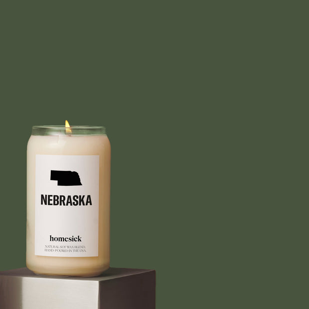 A lit Nebraska candle displayed on a silver cube in the front left corner of the image surrounding in a forest green studio enviornment.